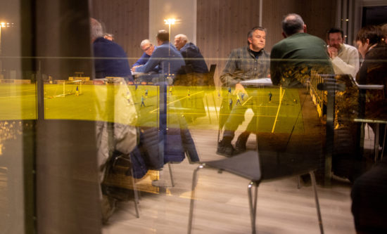 Mange personer samlet til gruppearbeid rundt mange bord. Fotballkamp speglar seg i vindauget bildet er tatt inn gjennom. Foto.