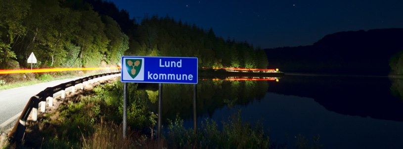 Skilt med "Lund Kommune". Mørkt, opplst av bil-lys. Foto.
