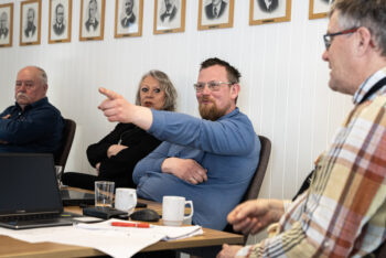 En mann i blå genser sitter i et møterom og peker mot noen utenfor bildet. Han smiler lurt. Tre andre personer sitter rundt han.