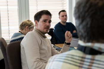 Fire personer sitter rundt et bord på et møterom. Mannen i midten av bildet ser mot en av de andre.