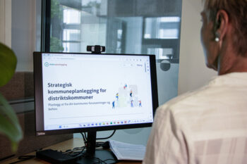 En kvinne står foran en kontorpult. PC-skjermen på pulten viser nettsiden enkelplanlegging.no
