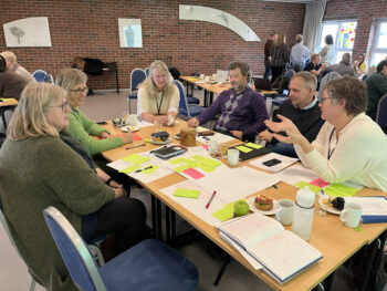 En gruppe som sitter rundt et bord og gjør gruppearbeid. Mange papirer, grønne og rosa lapper på bordet.