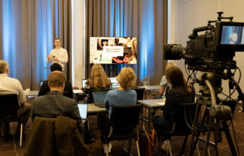 En mann holder en presentasjon. Han gestikulerer med hendene foran et publikum. Vi ser bakhodet på 7 personer i salen. I forgrunnen er et kamera som filmer mannen som holder presentasjon. Vi ser også powerpointpresentasjonen hans på en TV skjerm ved siden av foredragsholderen. 