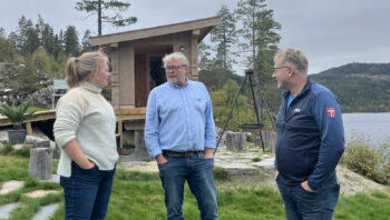 Tre personer står foran en badstue. Det er fjell, skog og vann bak personene og badstuen.