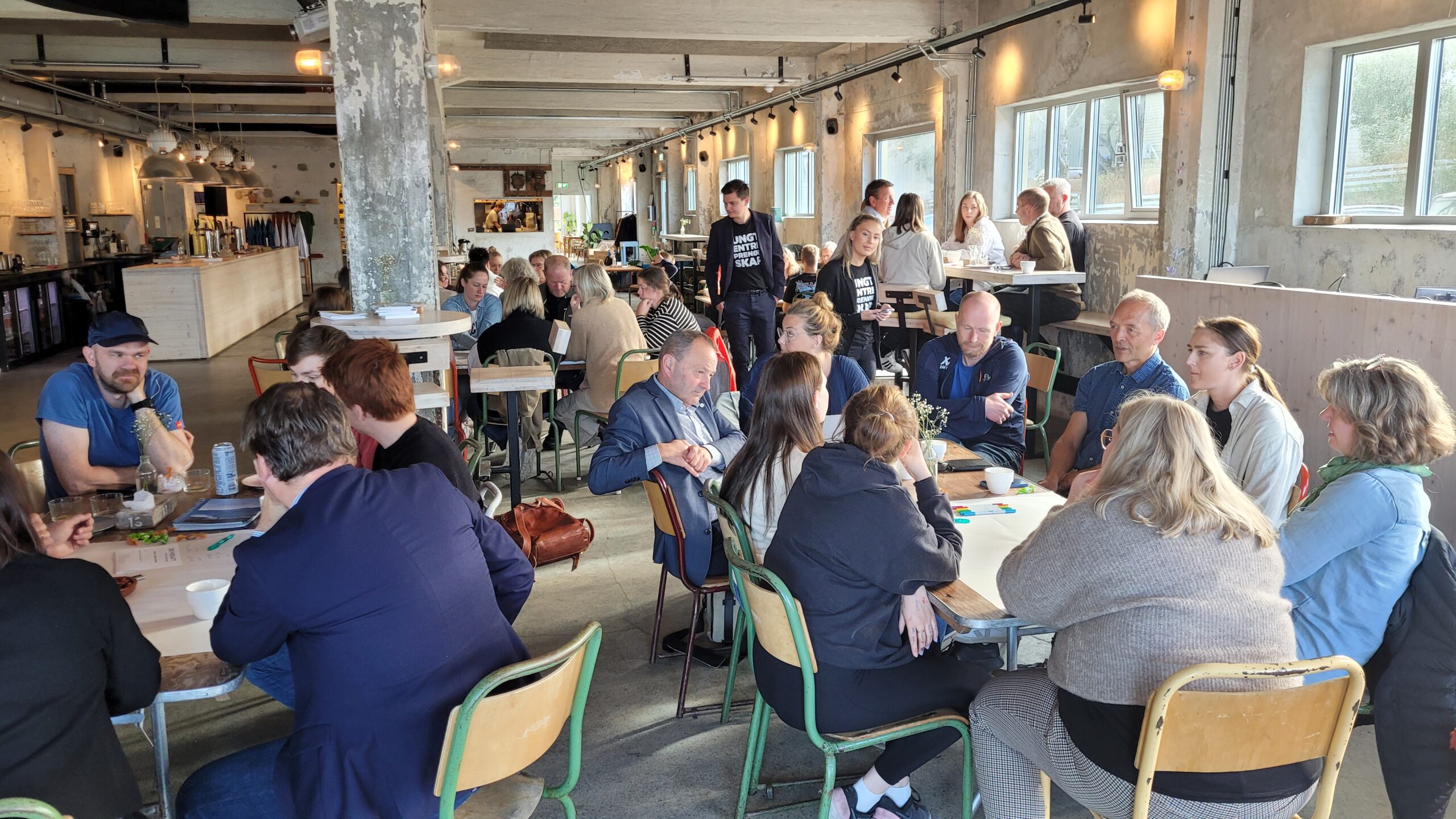 Et stort lokale med ca. 30 mennesker som sitter rundt bord og samarbeider.