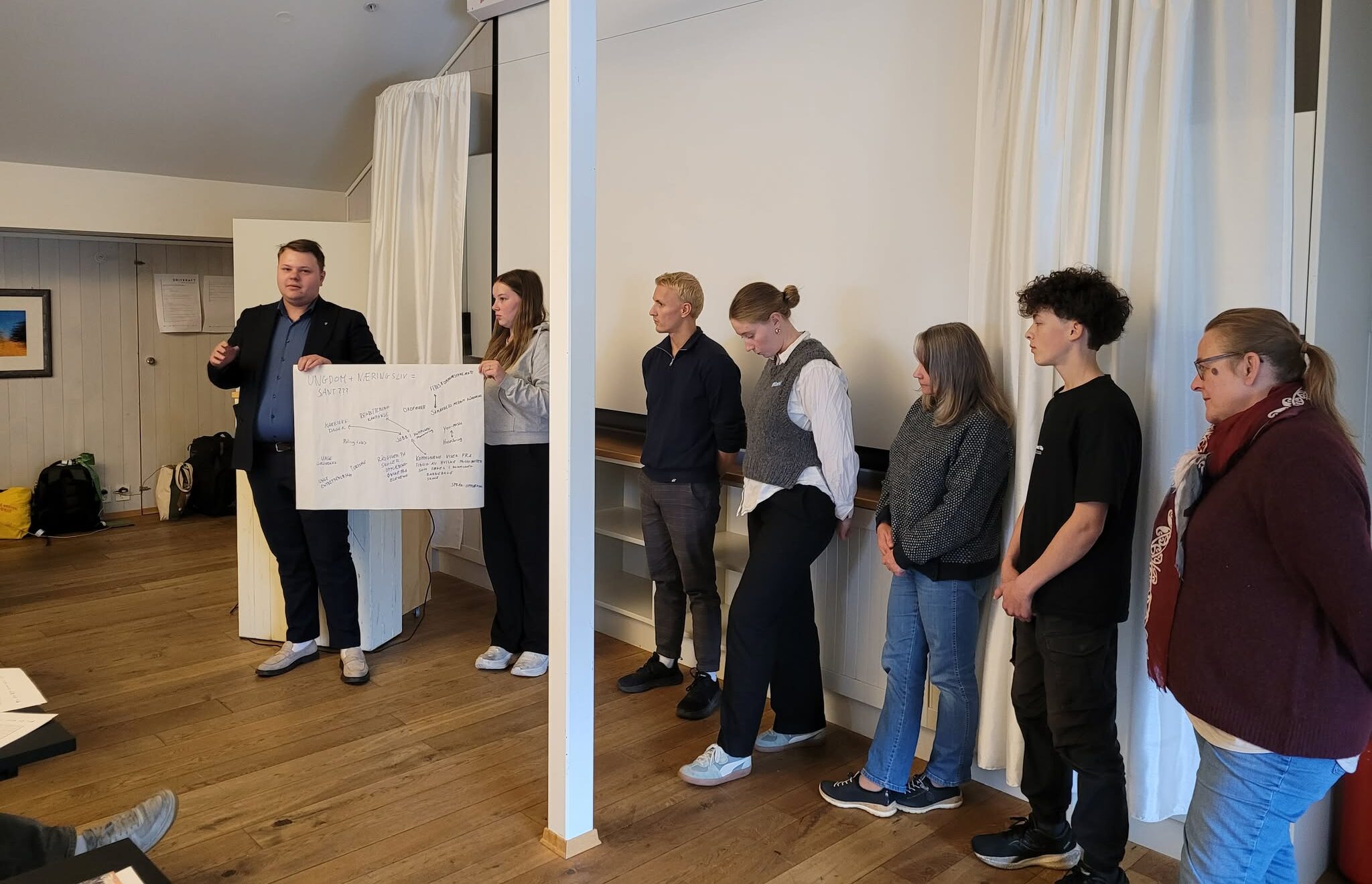 7 personer holder et innlegg. De står foran en storskjerm som er slått av. De fleste er ungdom. De to ungdommene som står helt til venstre holder opp en plakat. Det er ikke tydelig hva som står på plakaten.